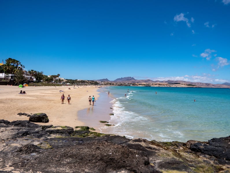 Langer Sandstrand mit Spaziergängern, klares blaues Meer, an der grünen Promenade reihen sich Häuser aneinander auf Fuerteventura