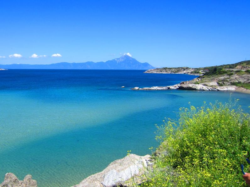 Blick über eine weite Bucht mit blauem Wasser und Bergen im Hintergrund.