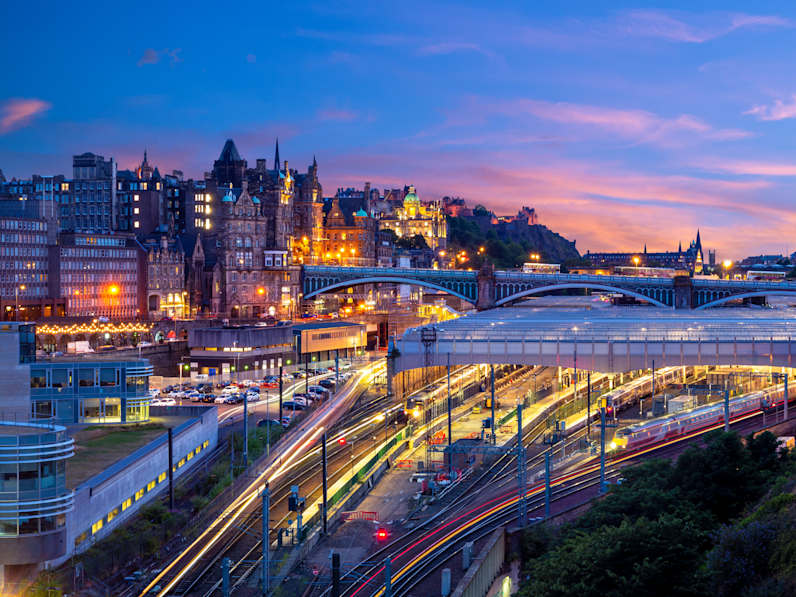 Die Gleise und das Bahnhofsgebäude in Edinburgh von oben. Dahinter erhebt sich ein kleiner Hügel, der mit  Häusern bebaut ist.