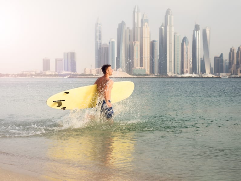 Surfer läuft ins Wasser vor der Hochhaus-Kulisse von Dubai.