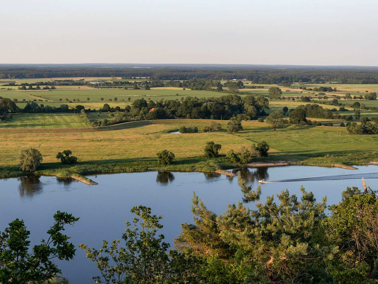 Blick über eine grüne flache Landschaft. Im Vordergrund fließt ein breiter Fluss, auf dem ein kleines Boot fährt.