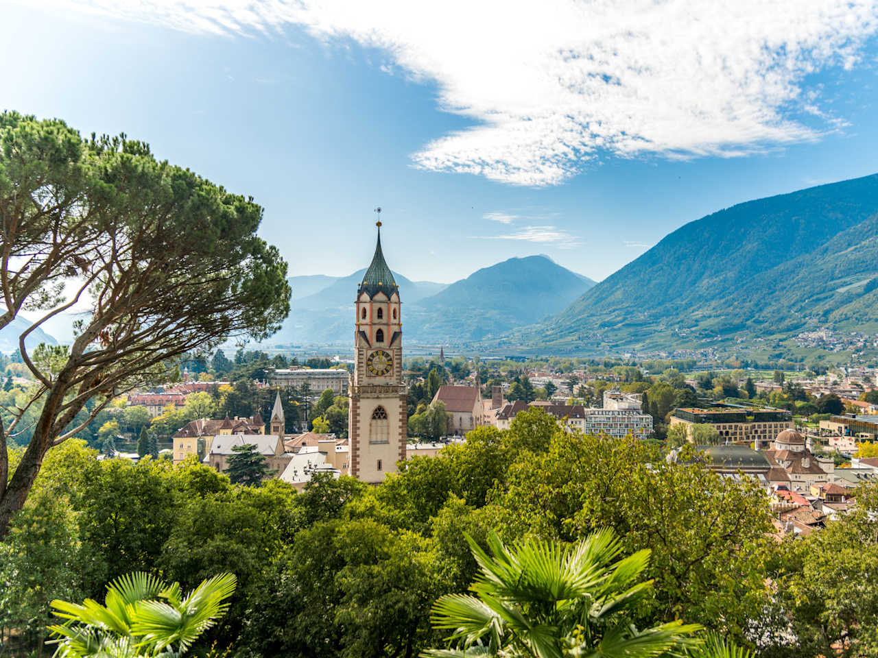 Der Kirchturm von St. Nikolaus überragt das grüne Stadtbild Merans – umgeben von Laubengängen, Promenaden und Bergen.