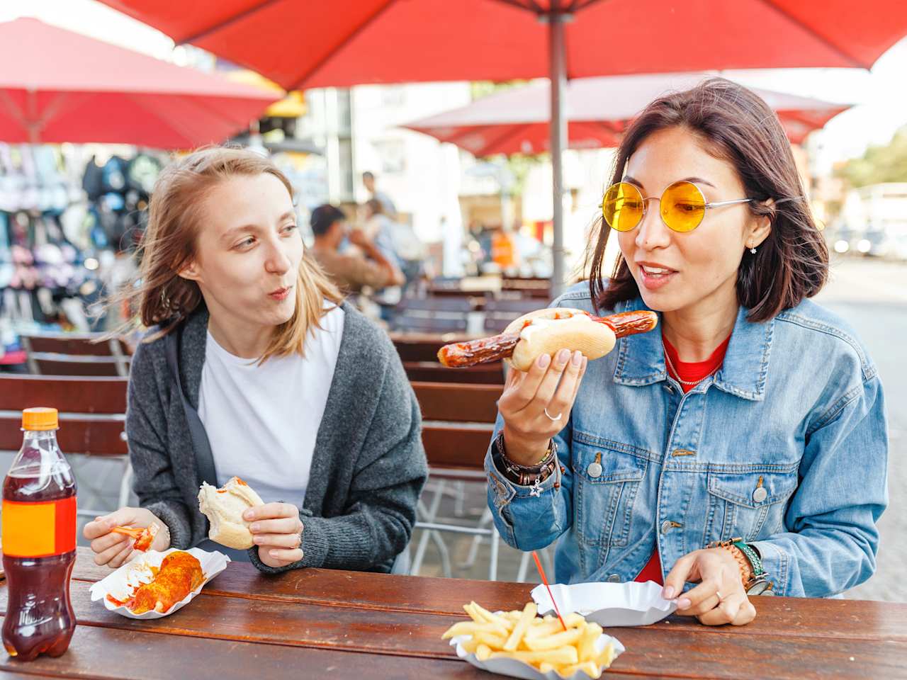 Zwei Frauen sitzen an einem Tisch unter roten Schirmen an einem Imbiss, die eine isst Currywurst mit Brötchen und die andere Bratwurst mit Pommes.