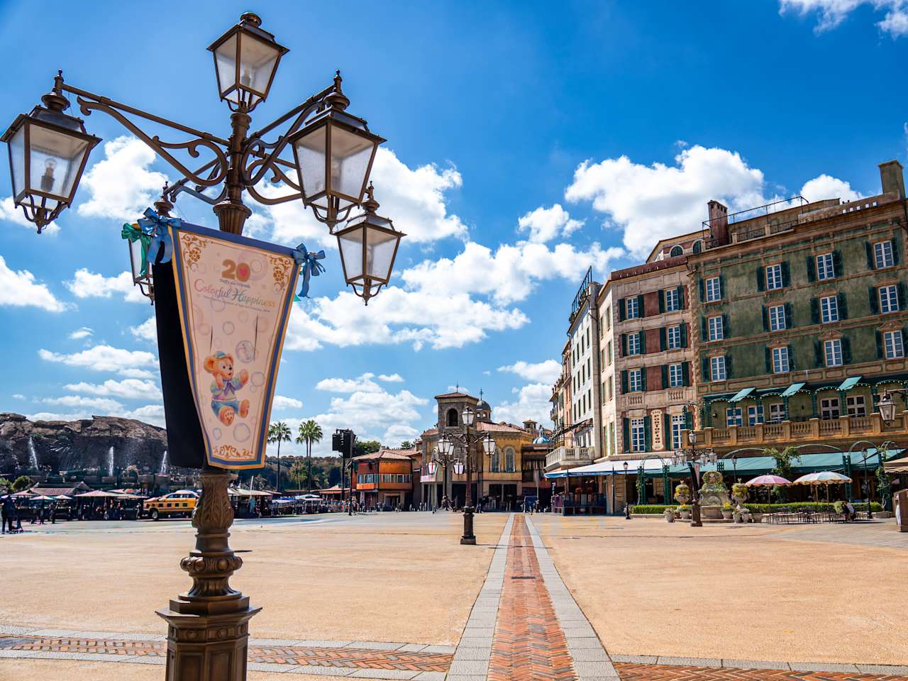 Ein Platz mit bunten, ästhetischen Häusern am Rand, einer verschnörkelten Laterne im Vordergrund und im Hintergrund Berge mit kleinen Wasserfällen im DisneySea bei Tokyo, Japan.