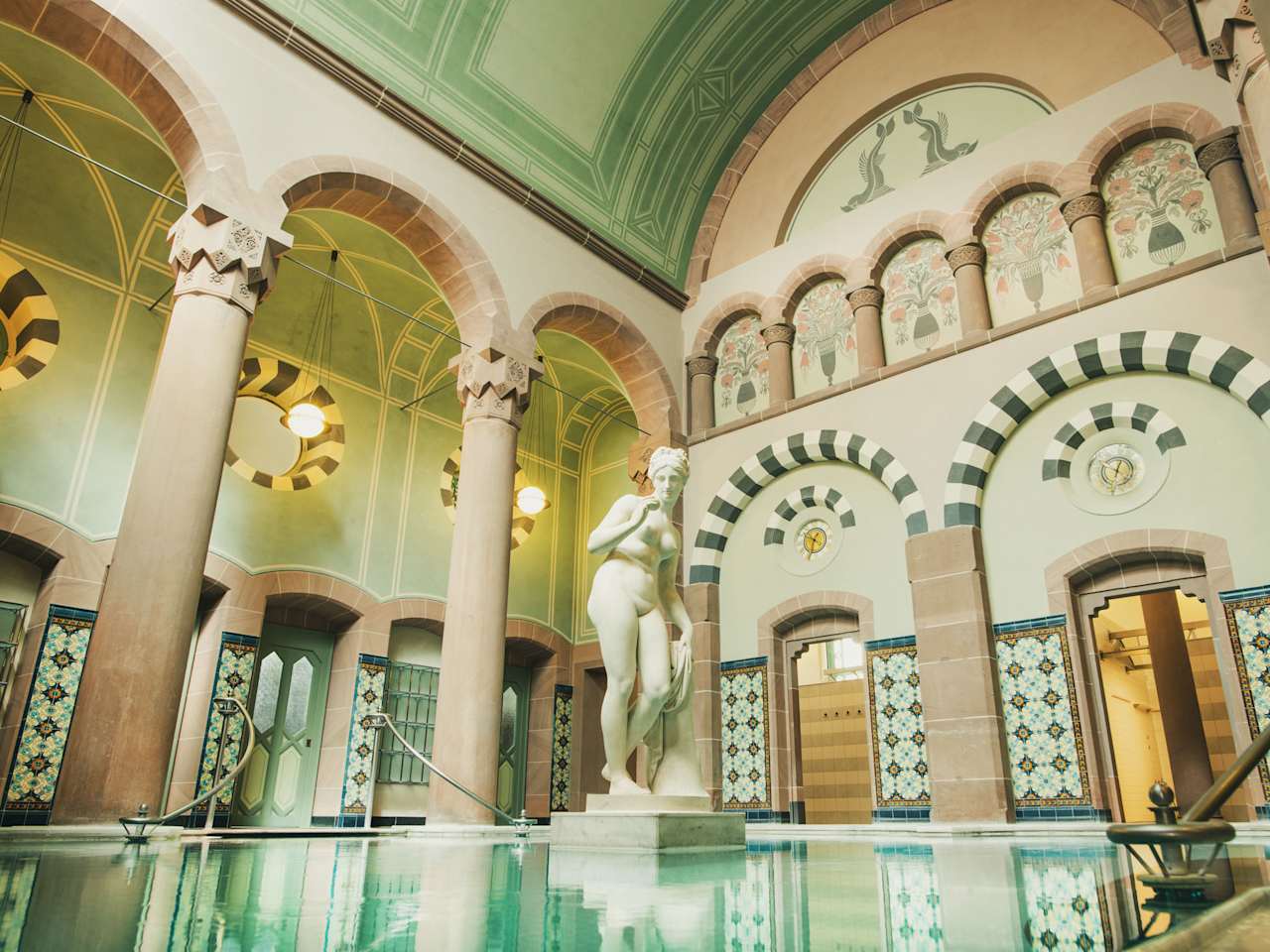 Eleganter Thermalraum mit großer Marmorstatue in einem Becken, umgeben von filigran bemalten Rundbögen, hohen Säulen, Jugendstil-Ornamentik und farbigen Wandfliesen im Palais Thermal Bad Wildbad.