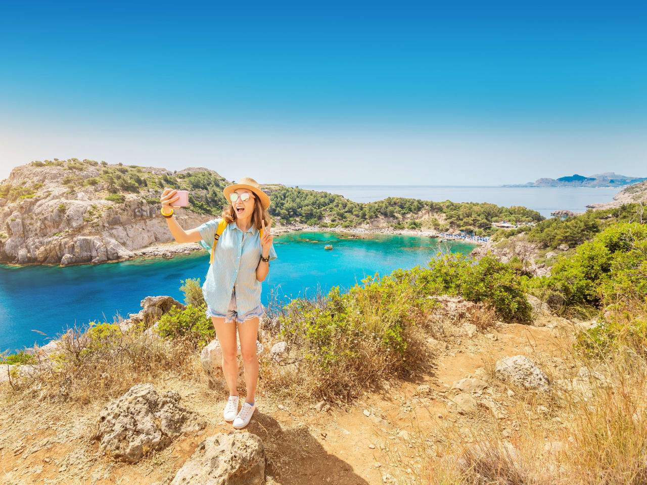 Mädchen mit Hut macht ein Selfie mit einer azurblauen Mittelmeer-Bucht im Hintergund © iStock.com/frantic00