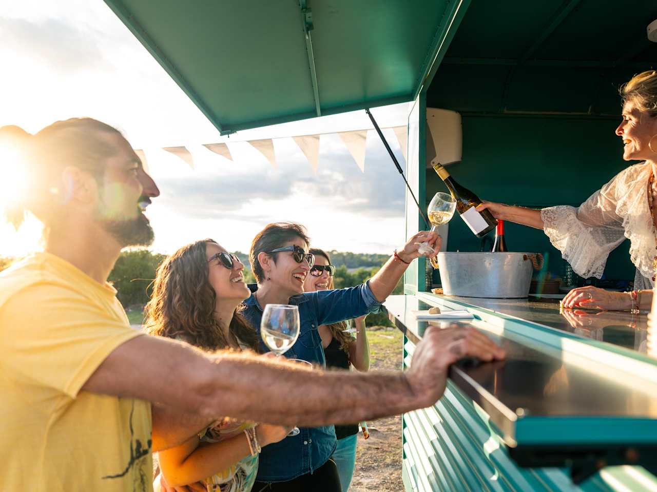 Vier Menschen stehen vor einem blau-grünen Foodtruck und halten Weingläser in der Hand. Im Truck steht eine Frau und schenkt Weißwein aus.