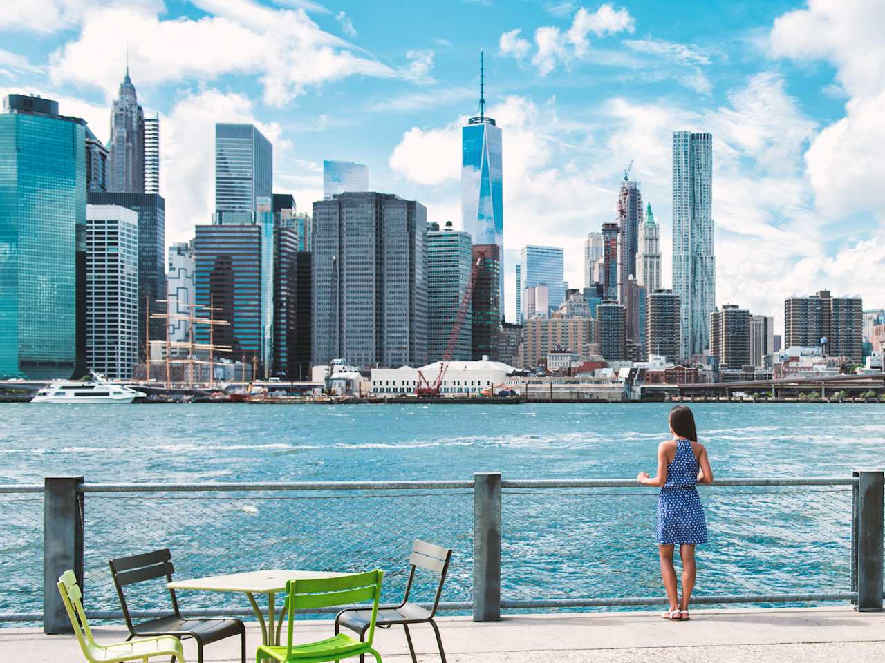Eine Frau in einem blauen Kleid steht an einem Brückenzaun. Auf der anderen Seite liegt ein Fluss und eine Skyline.
