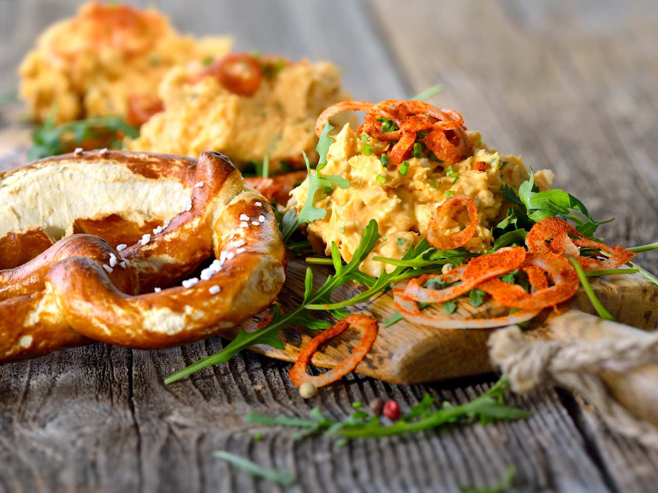 Ein kleines Holzbrett voller Obatzter auf Rucola liegt neben einer „Brezn“.