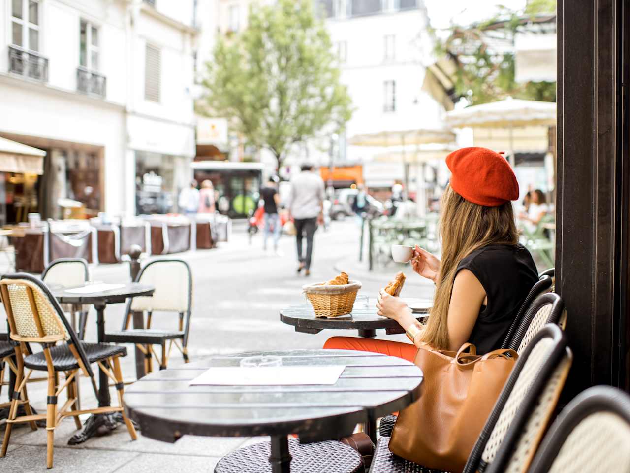 Eine Frau mit rotem Beret sitzt an einem Cafétisch und hält ein Croissant und einen Espresso in den Händen.