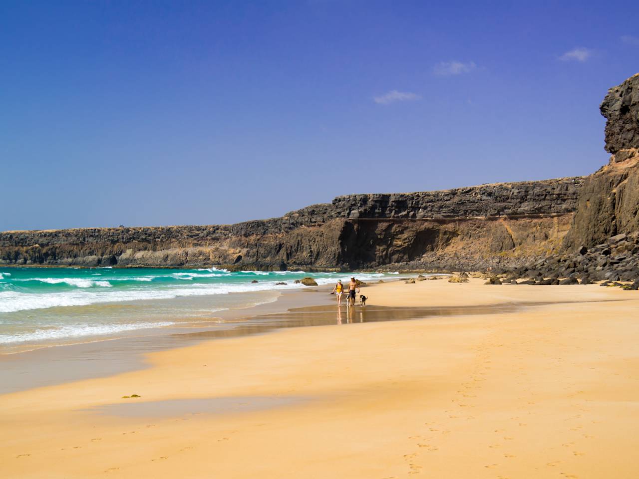 Blick auf die Naturstrand-Bucht Playa del Águila, Fuerteventura mit Steilküste und zwei Menschen mit Hund am Strand
