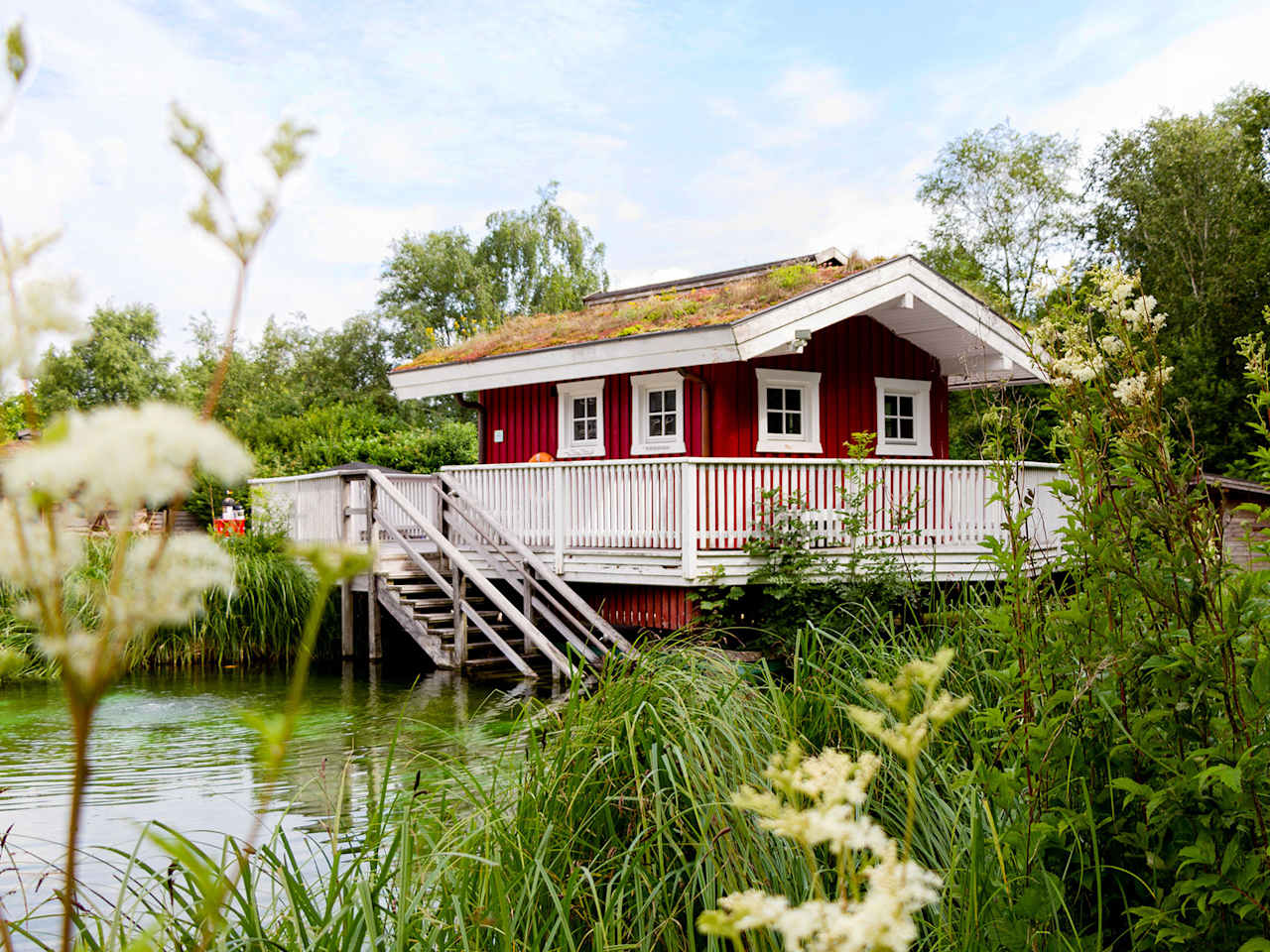 Rote skandinavisch anmutende Saunahütte mit Gründach und Holzterrasse, auf Stelzen über einem natürlichen Teich gelegen, umgeben von grüner Vegetation in der Friesentherme Emden.