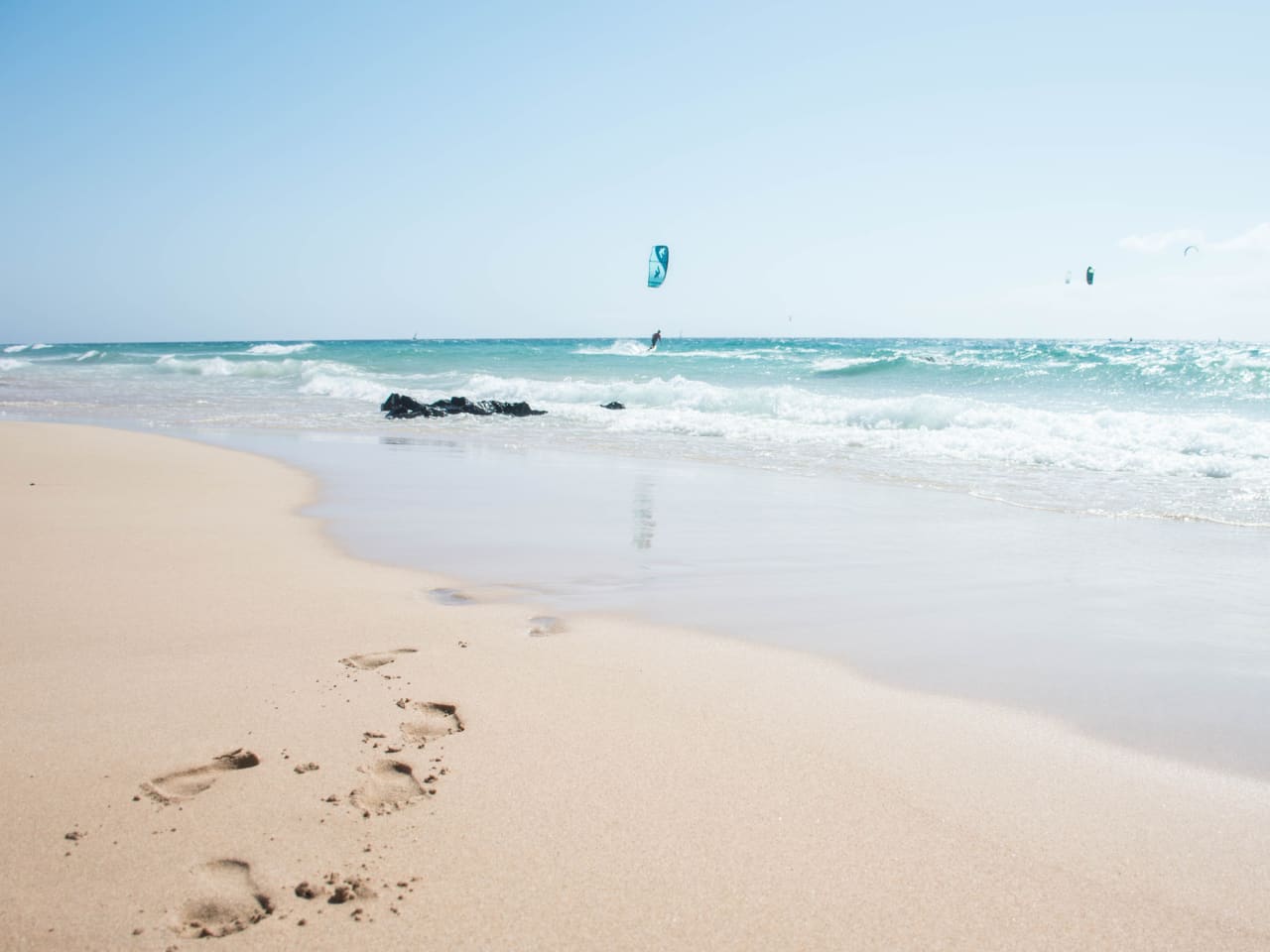 Sandstrand und blaues Meer mit Fußabdrücken und Menschen die kiteboarden © iStock.com/Pia Staric