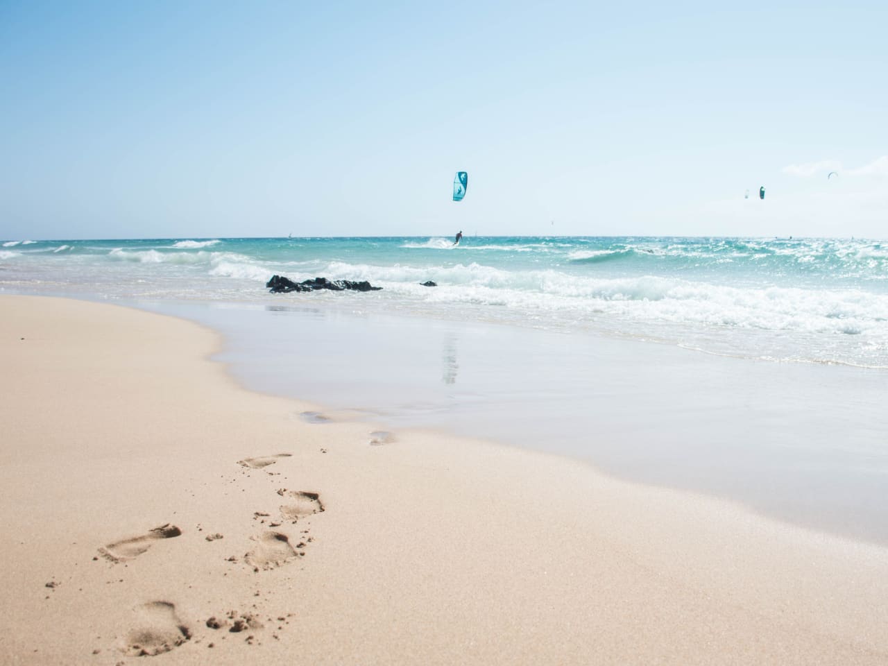 Sandstrand und blaues Meer mit Fußabdrücken und Menschen die kiteboarden © iStock.com/Pia Staric