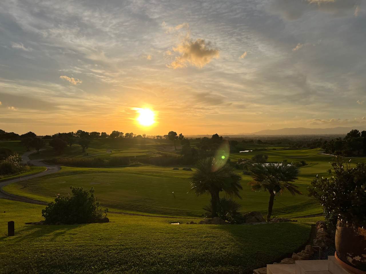 Ein weitläufiger, gepflegter Golfplatz mit Palmen und hügeligem Grün wird in warmes Licht eines Sonnenuntergangs getaucht, im Hintergrund Berge und weite Landschaft bei Palma de Mallorca.