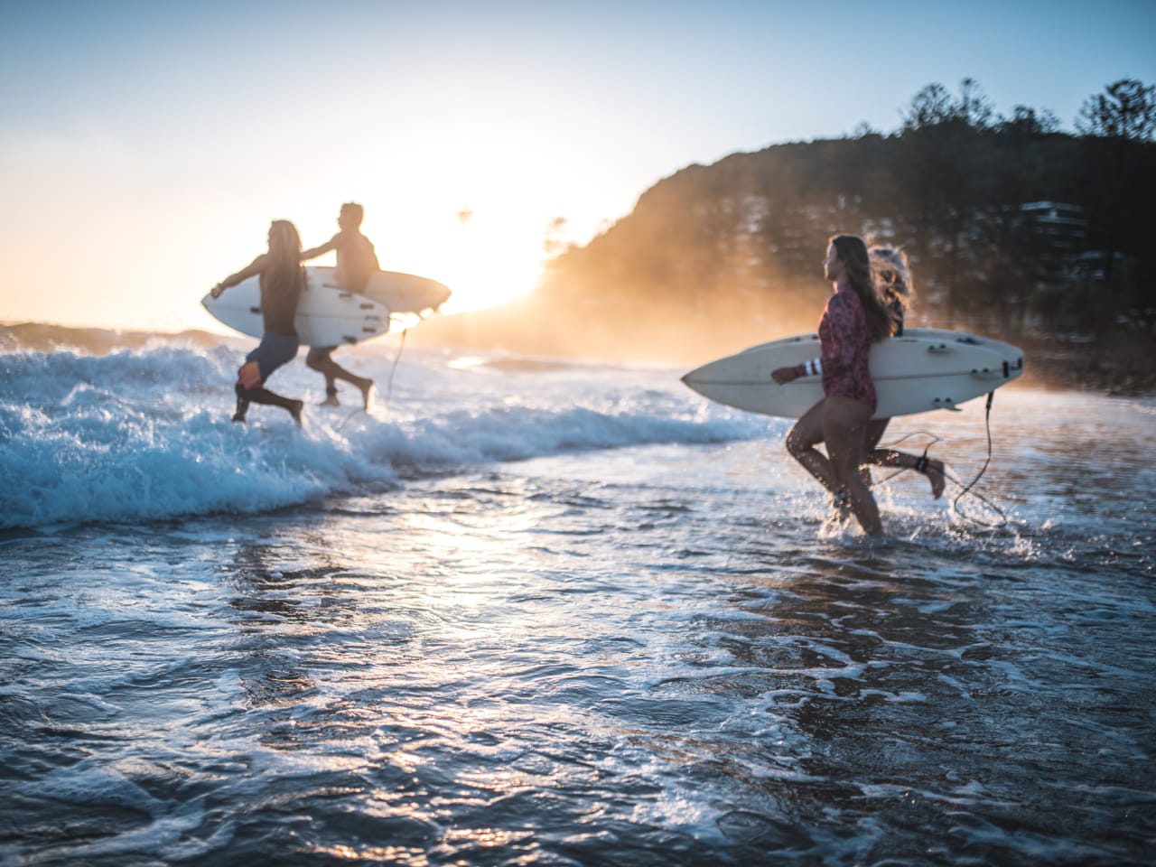 Vier Freunde laufen früh morgens mit Surfbrettern in der Hand ins Wasser © iStock.com/AzmanL