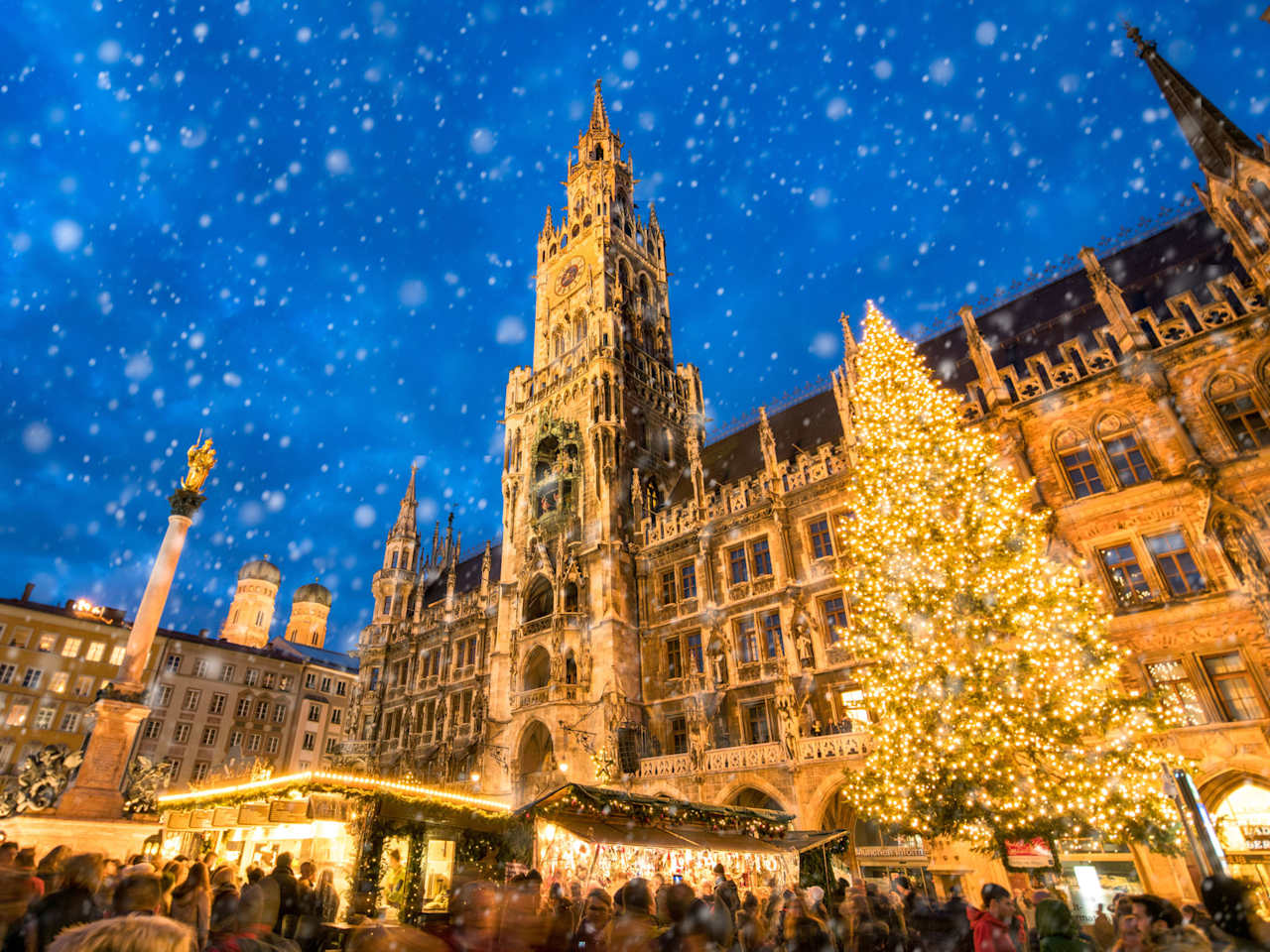 Winterliche abendliche Szene des Christkindlmarkts am Marienplatz in München, mit dem beleuchteten Rathaus, einem großen geschmückten Weihnachtsbaum und Schneefall über den Marktständen.