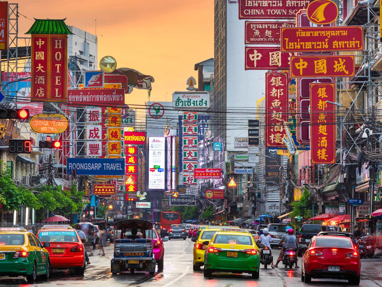 Autos fahren zwischen Hochhäusern entlang. Überall sind Werbeschilder mit thailändischer, chinesischer und englischer BEschriftung zu sehen.