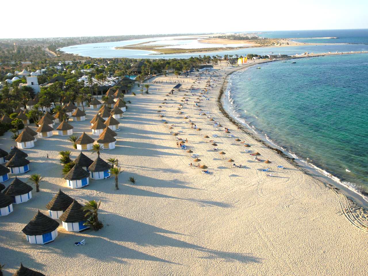 Drohnenaufnahme von einem hübschen Strand und dem blauen Meer. Auf dem Strand stehen einige Hütten, Sonnenschirme und Palmen.