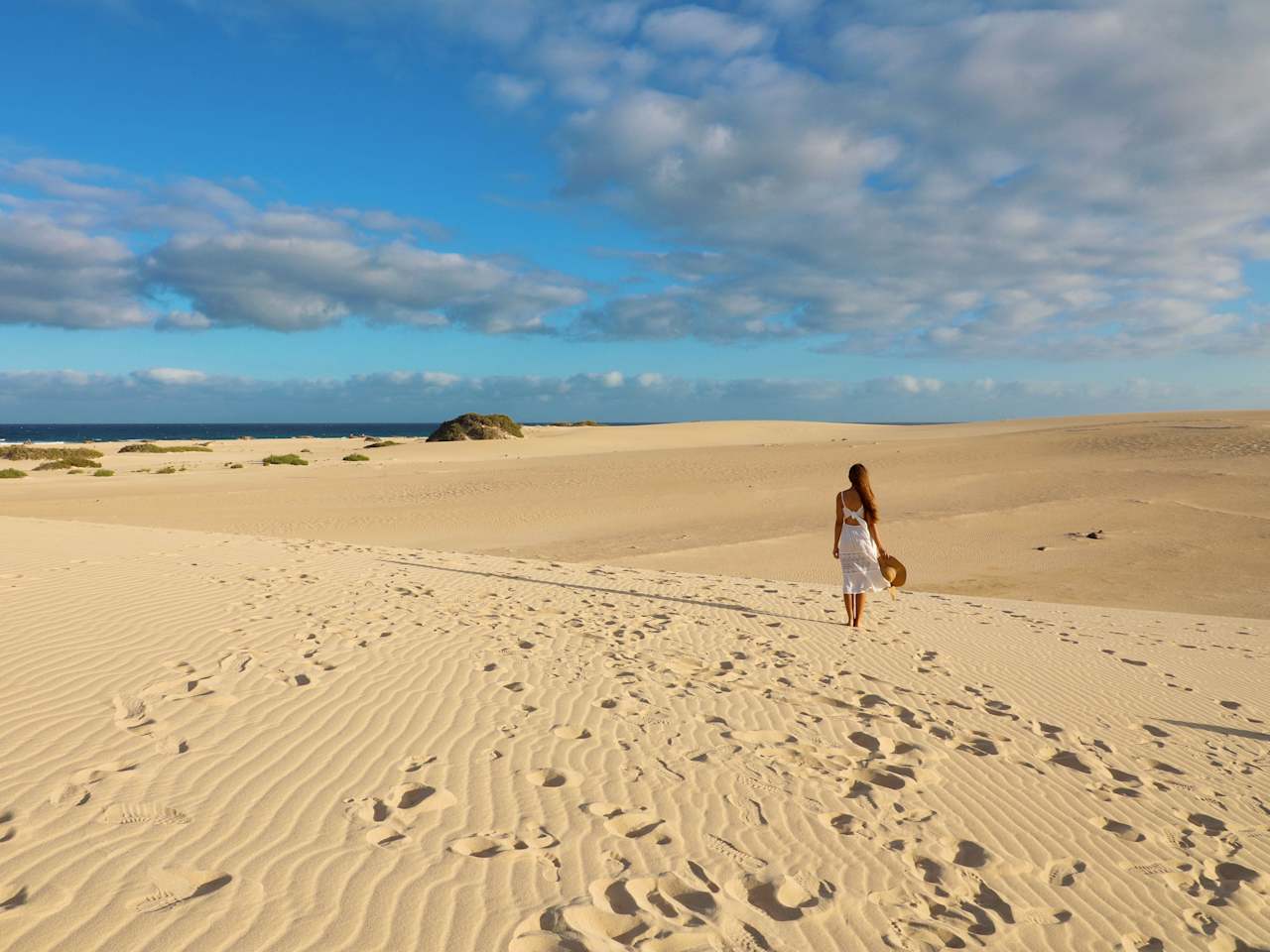 Eine Frau läuft im weißen Sommerkleid und mit Hut in der Hand über die Dünen bei Corralejo, Fuerteventura