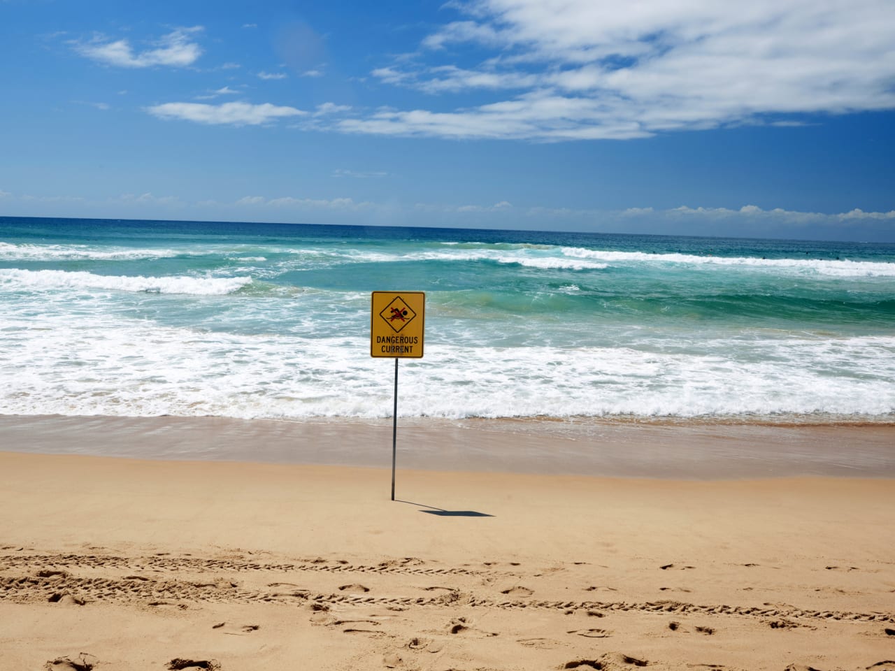 Ein gelbes Warnschild an einem Strand direkt am Meer.