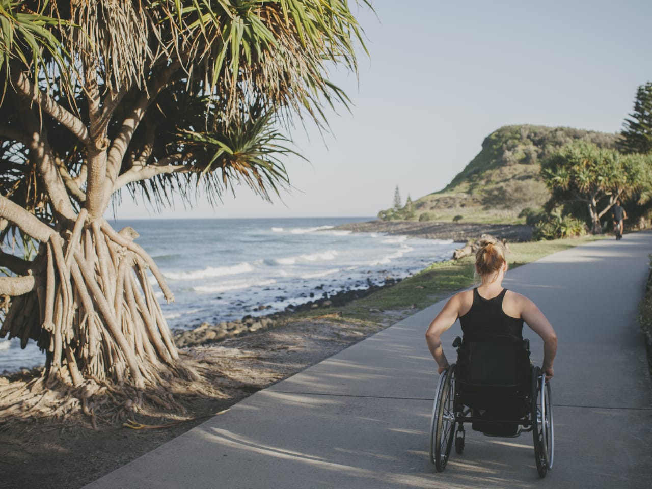 Eine Frau fährt in einem Rollstuhl auf einem Weg direkt am Meer.