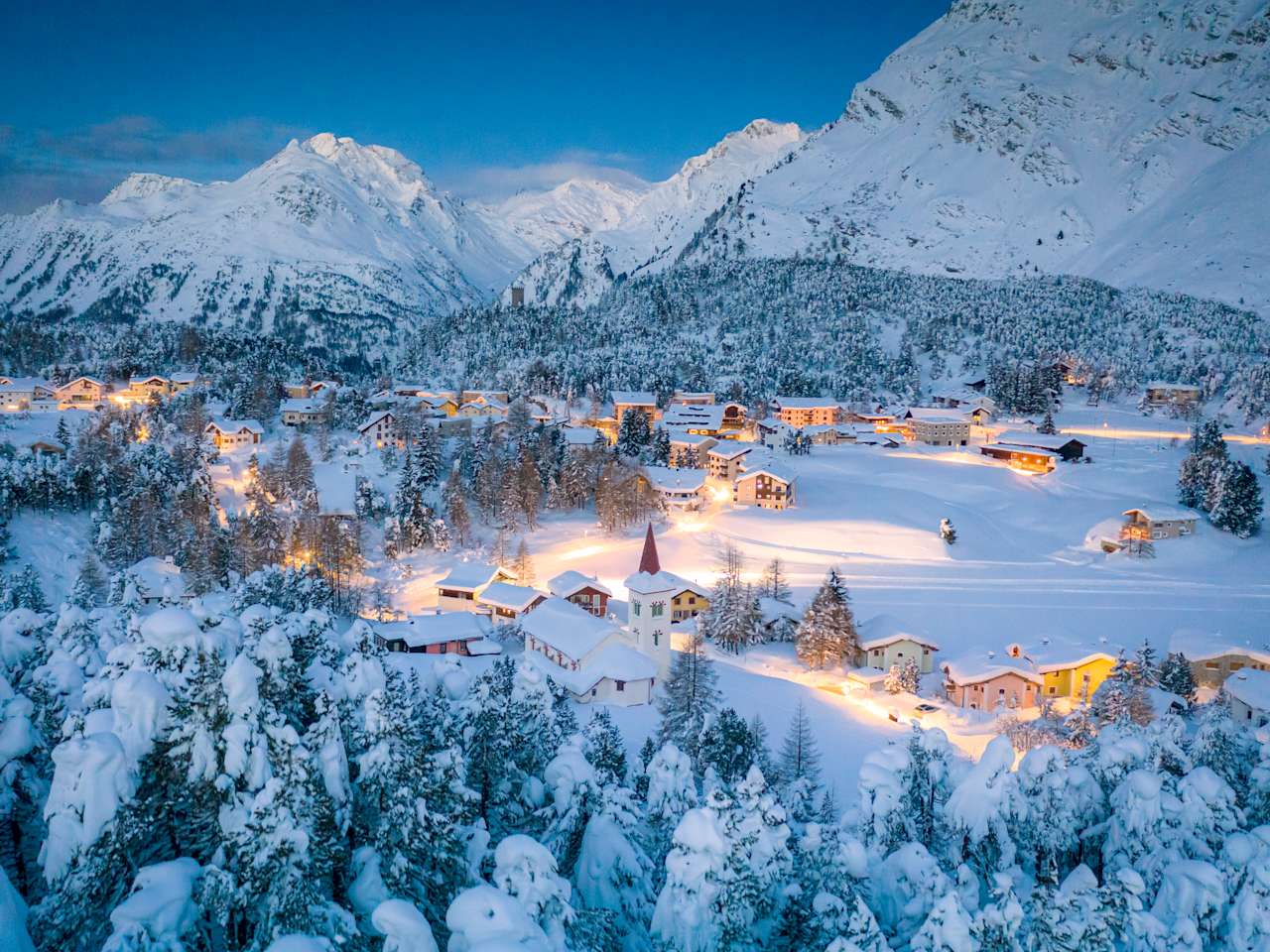 Alpendorf Maloja und Chiesa Bianca umrahmt von verschneiten Wäldern in der Abenddämmerung, Bergell, Kanton Graubünden, Engadin, Schweiz. © Roberto Moiola / Sysaworld via Getty Images