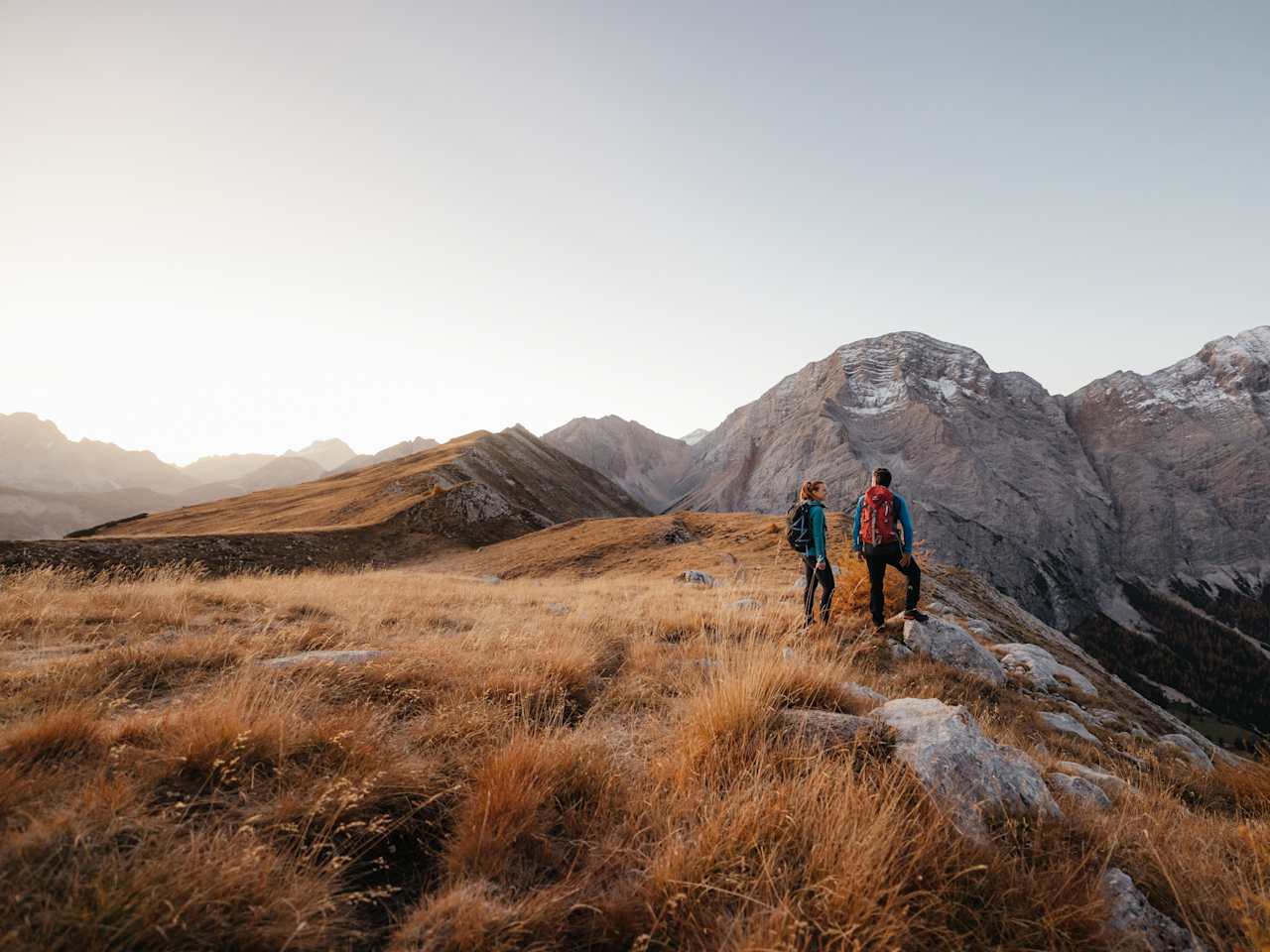 Zwei Wandernde mit Rucksäcken stehen auf einem schmalen Berggrat und blicken auf felsige Dolomitengipfel im warmen Licht