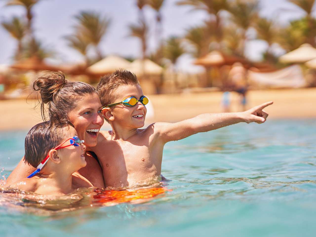 Eine Mutter ist mit ihren zwei Kindern im Wasser und hält sie im Arm. Im Hintergrund sieht man verschwommen den Strand und Palmen