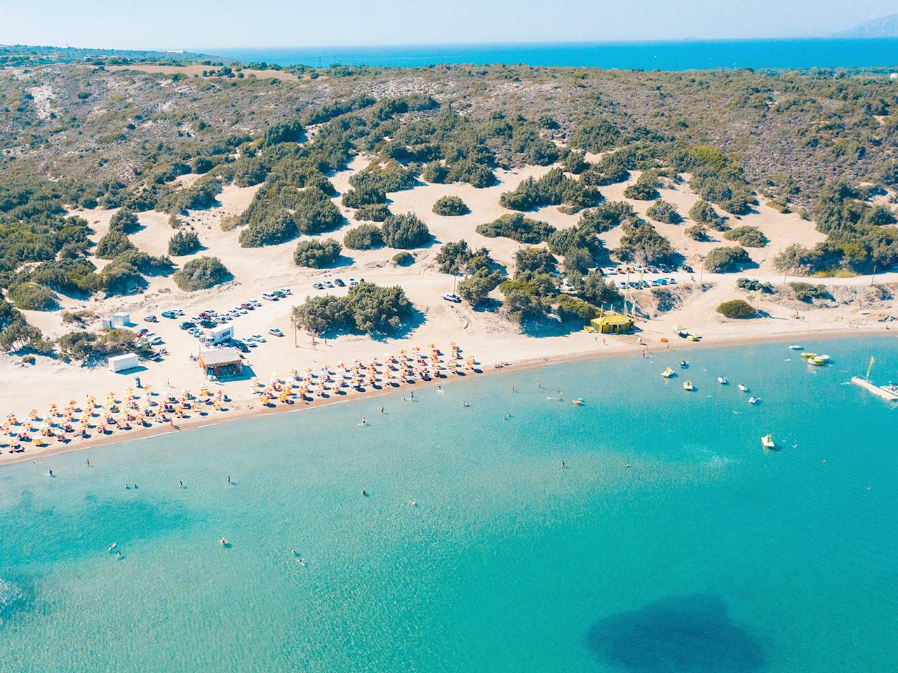 Ein weißer Sandstrand mit Sonnenschirmen grenzt an das klare Meer. Im Hintergrund sind viele Sträucher zu sehen, die auf Dünen wachsen.