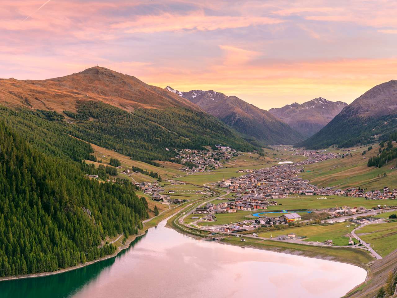 Blick über einen See auf den Ort Livigno in einem breiten Alpental bei Sonnenuntergang, umgeben von Bergen und Wäldern.