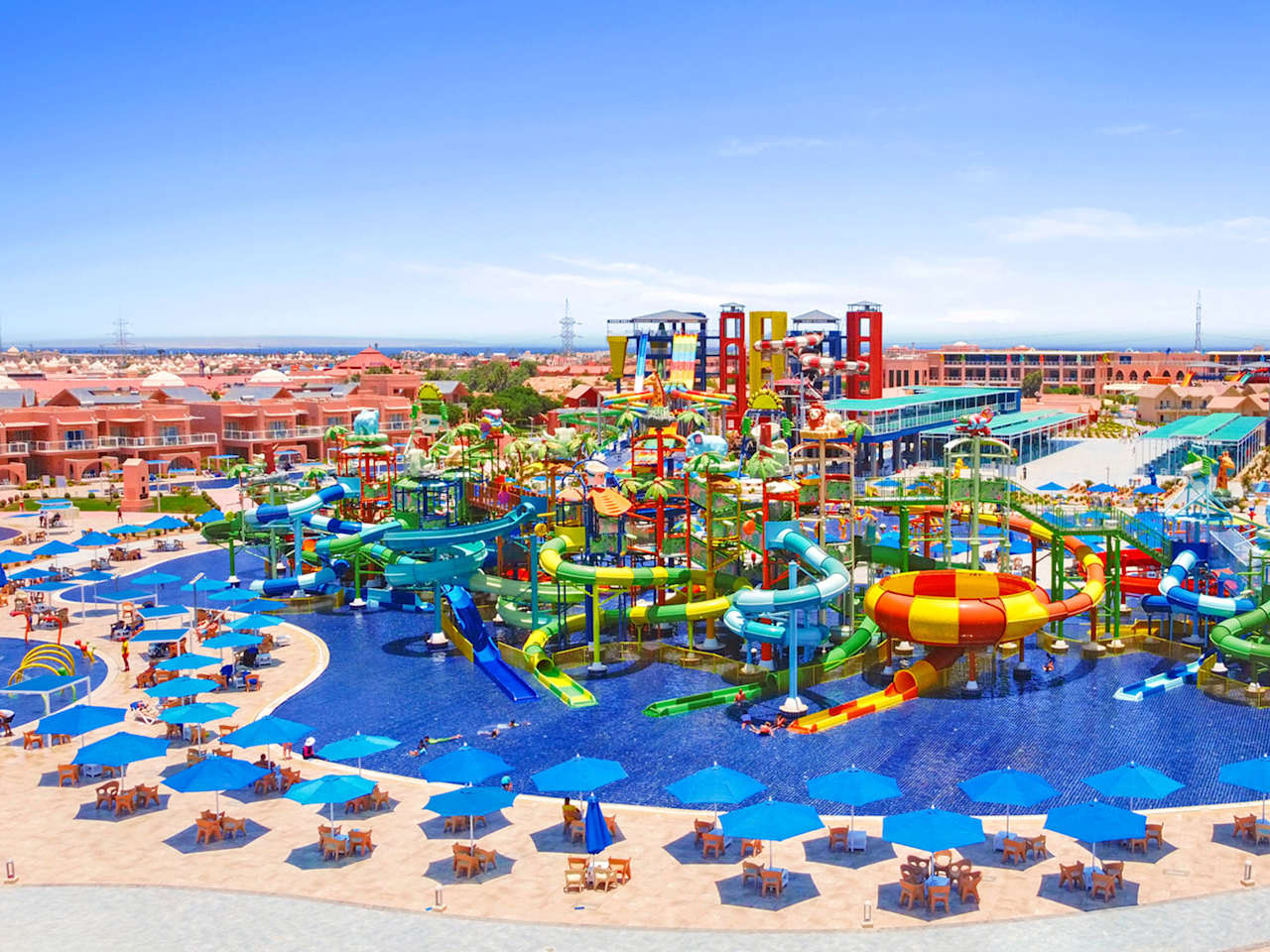 Luftaufnahme eines Wasserparks mit grünen, gelben und roten Rutschen, großem Poolbereich und zahlreichen blauen Sonnenschirmen rund um das Becken.