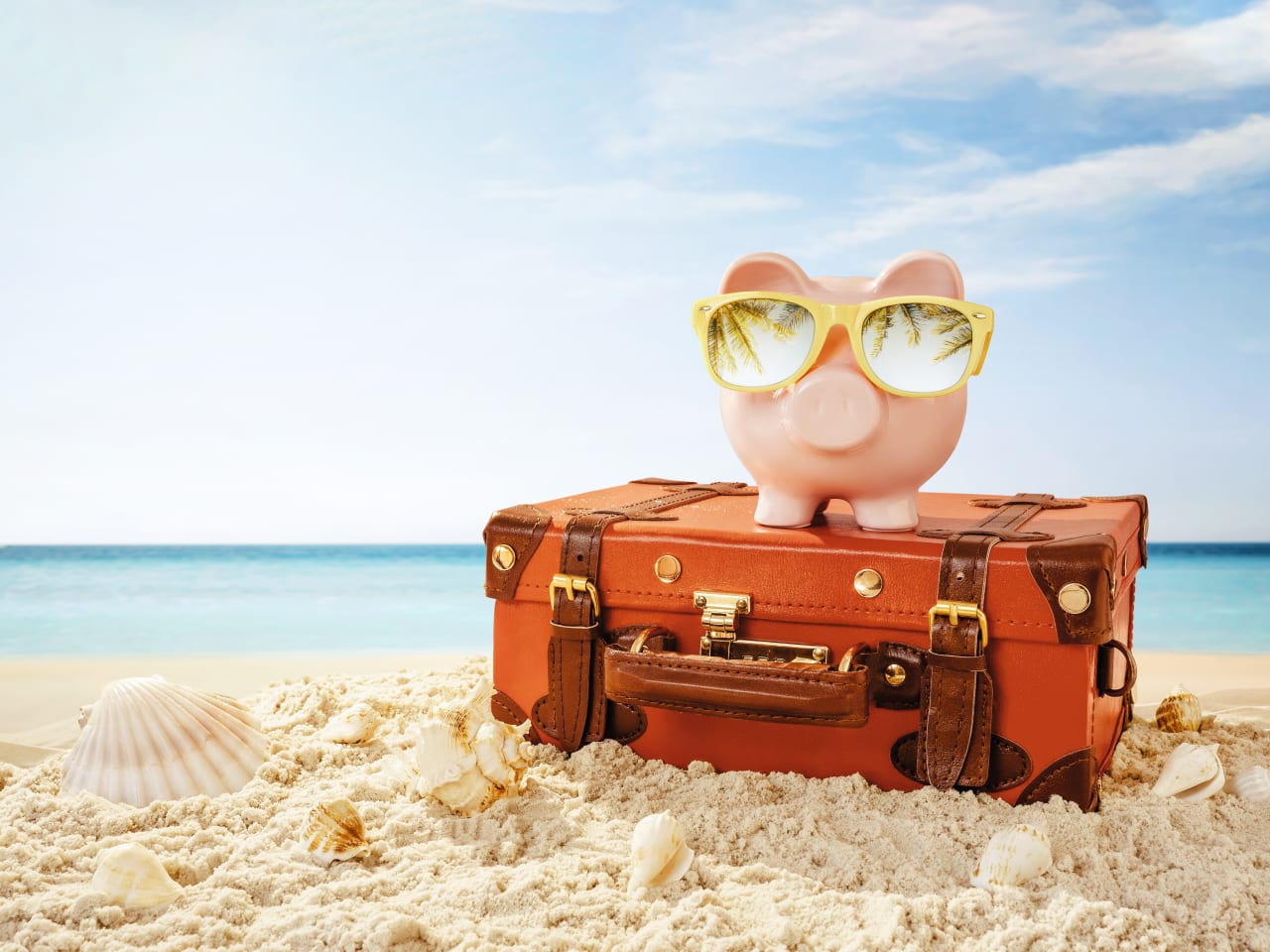 Sparschwein mit Sonnenbrille und Reisekoffer am Strand
