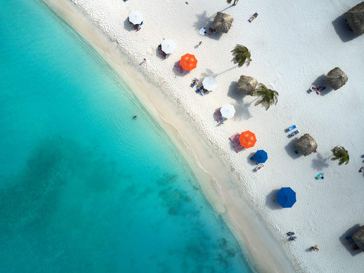Ansicht von oben. Links ist das Meer, rechts ein Strand mit Sonnenschirmen, Palmen und Strandliegen.
