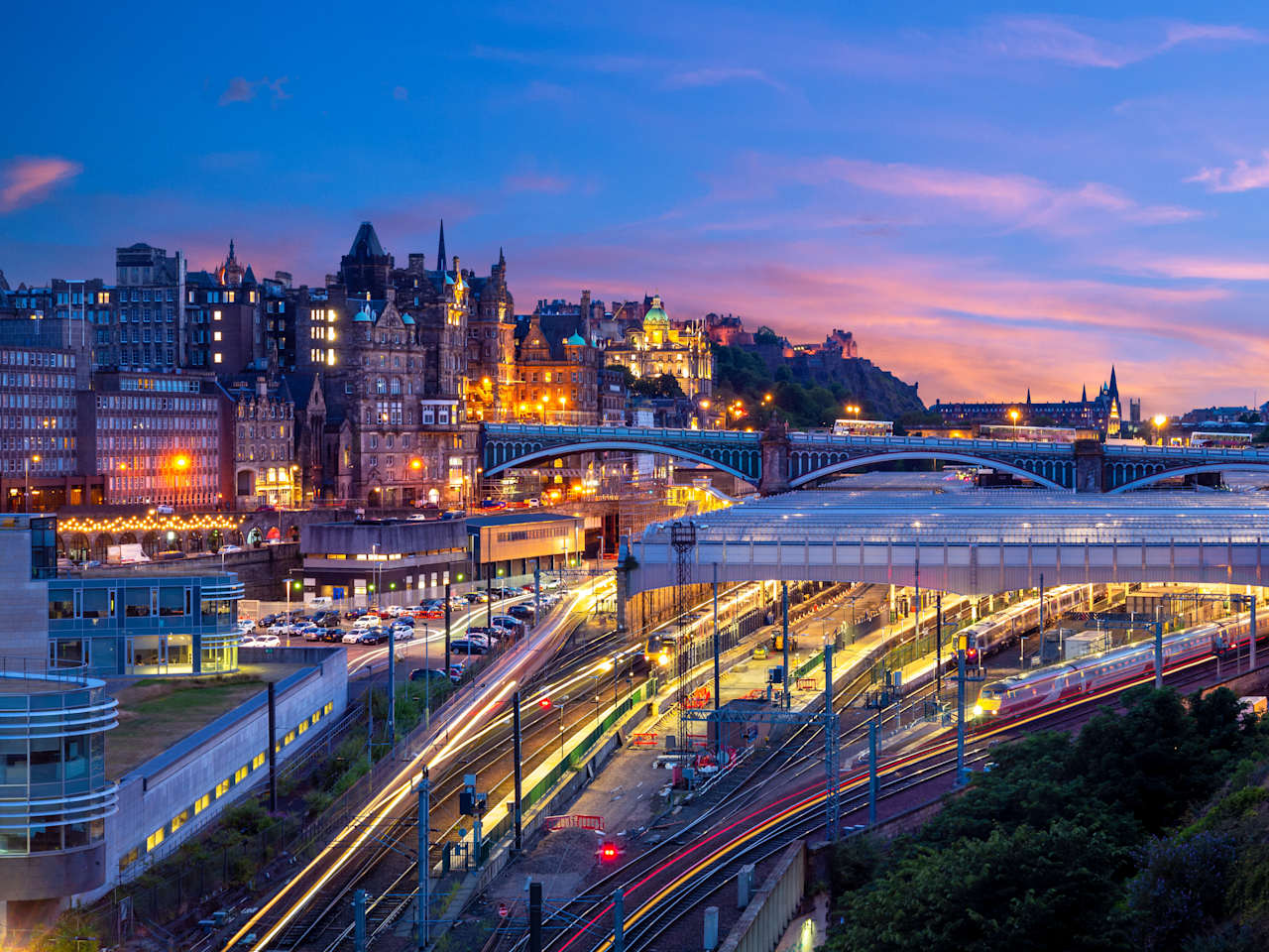 Die Gleise und das Bahnhofsgebäude in Edinburgh von oben. Dahinter erhebt sich ein kleiner Hügel, der mit  Häusern bebaut ist.