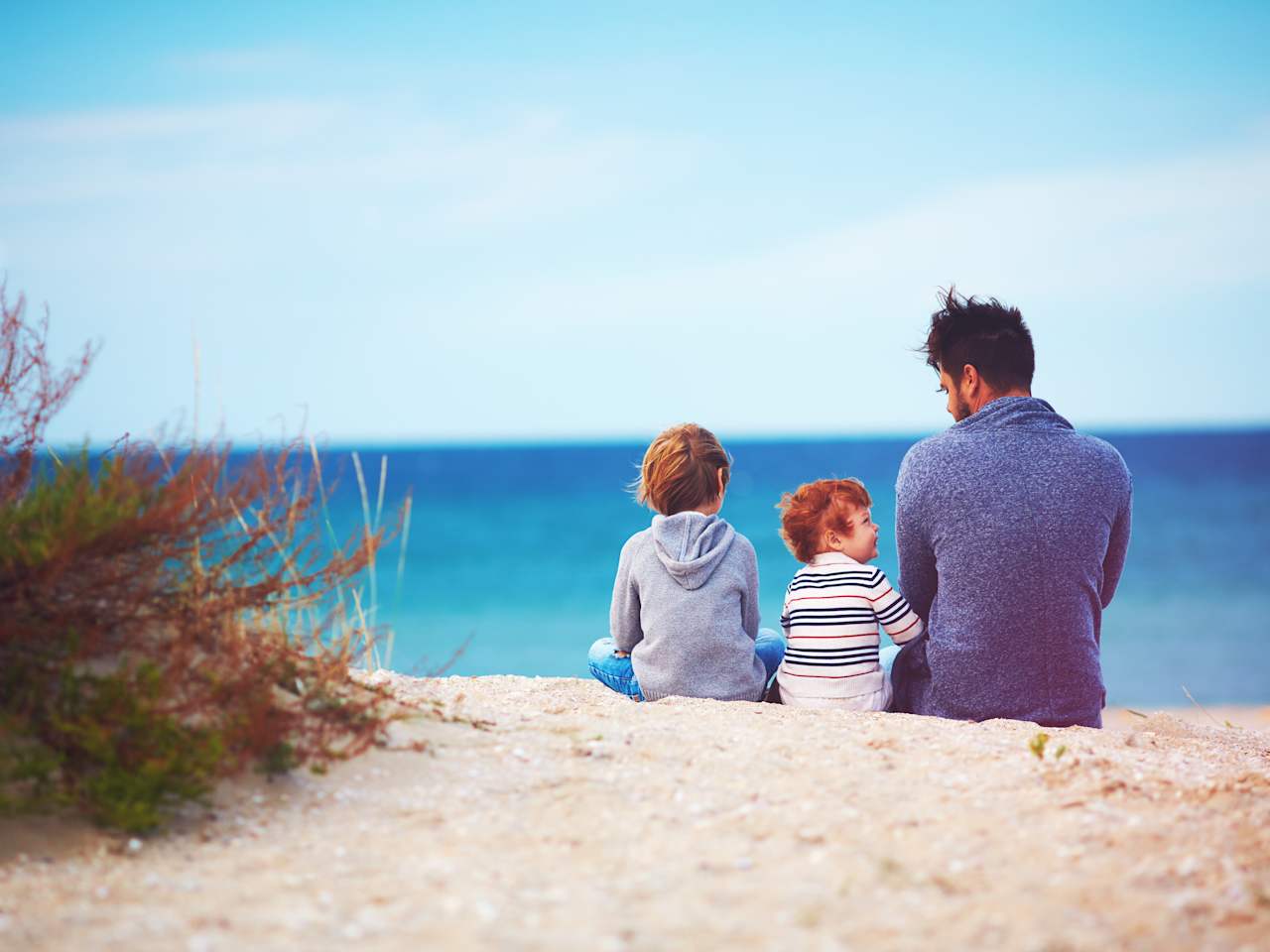 Vater sitzt mit seinen beiden kleinen Söhnen im Sand am Meer.
