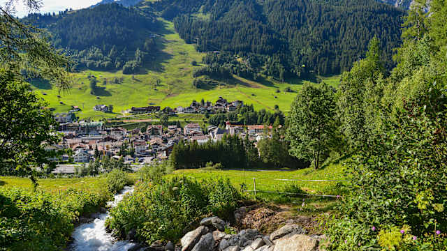 Blick aus einem kleinen Wald auf ein Dorf. Zu Füßen fließt ein Fluss bergab Richtung Dorf. Dahinter leigt ein grüner Berg.