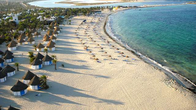 Drohnenaufnahme von einem hübschen Strand und dem blauen Meer. Auf dem Strand stehen einige Hütten, Sonnenschirme und Palmen.
