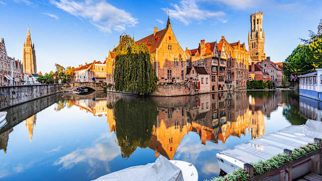 Ein mittelalterliches Bauensemlbe mit mehreren Häusern und einem Turm spiegelt sich im Wasser eines Kanals.
