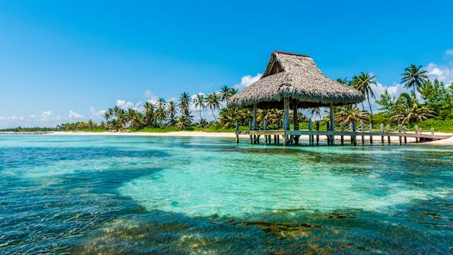 Blick auf eine Strandhütte am Strand mit Palmen und türkisblauem Wasser. © Marco Bottigelli via Getty Images