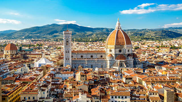 Blick über ein Häusermeer zwischen dem sich der große Dom von Florenz erhebt. Das weiße Gotteshaus ist mit blauen rechteckigen Formen verziert und hat – wie der Rest der Stadt – rote Dächer.