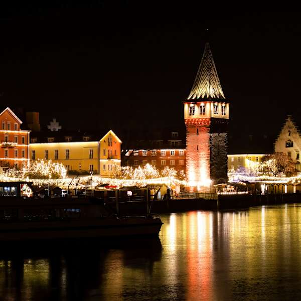 Nächtliche Ansicht der Lindauer Hafenweihnacht mit beleuchteten Häusern, Marktständen und dem angestrahlten Mangturm; die Lichter spiegeln sich deutlich im ruhigen Wasser des Bodensees.