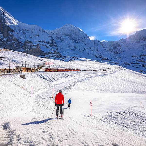Eine Bahn fährt durch eine verschneite Berglandschaft. Daneben fahren zwei Skifahrer den Berg hinab.